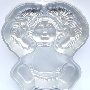 Könnte beinhalten: Eine silberne Kuchenform aus Metall in Form einer Puppe mit Zöpfen. Die Puppe hat ein rundes Gesicht mit großen Augen und einem Lächeln.