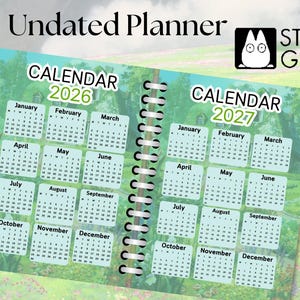 May include: An undated planner with a green and blue cover featuring a calendar for 2026 and 2027. The cover has a whimsical illustration of a forest scene with a house and a spiral binding.
