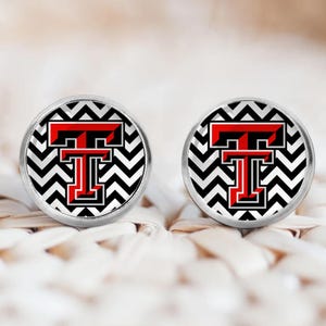 May include: A pair of round stud earrings. Each earring features a red and black letter "T" on a black and white chevron pattern background, set in a silver-colored metal frame. The earrings are displayed on a woven surface.