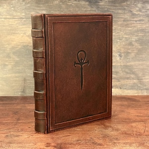 Puede incluir: Un libro encuadernado en cuero marr&oacute;n con un dise&ntilde;o en relieve en la portada. El lomo tiene bandas en relieve y la portada presenta un s&iacute;mbolo ankh estilizado. El libro est&aacute; de pie sobre una superficie de madera.