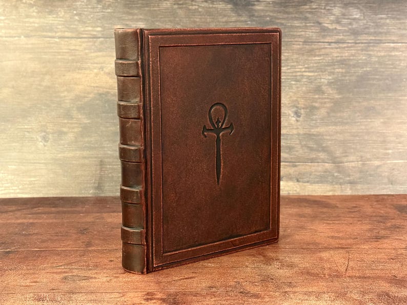 Puede incluir: Un libro encuadernado en cuero marr&oacute;n con un dise&ntilde;o en relieve en la portada. El lomo tiene bandas en relieve y la portada presenta un s&iacute;mbolo ankh estilizado. El libro est&aacute; de pie sobre una superficie de madera.