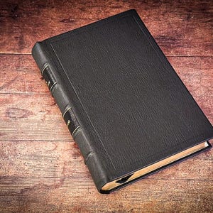 Puede incluir: Un libro negro de tapa dura con detalles dorados en el lomo. El libro tiene una cubierta texturizada y est&aacute; sobre una superficie de madera. Las p&aacute;ginas son visibles y tienen un tono dorado.