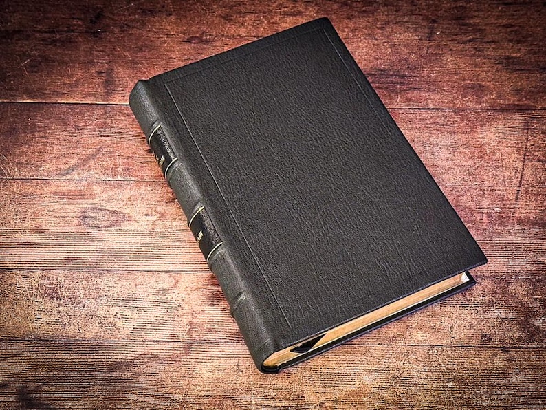 Puede incluir: Un libro negro de tapa dura con detalles dorados en el lomo. El libro tiene una cubierta texturizada y est&aacute; sobre una superficie de madera. Las p&aacute;ginas son visibles y tienen un tono dorado.