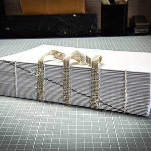 Puede incluir: Una pila de hojas de papel blancas encuadernadas con una cinta beige. Las p&aacute;ginas est&aacute;n cosidas con hilo de color claro. El libro est&aacute; sobre una superficie gris con un patr&oacute;n de cuadr&iacute;cula, lo que sugiere un proyecto de artesan&iacute;a o arte.