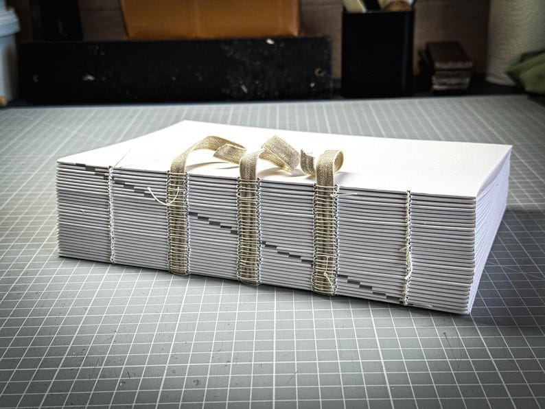 Puede incluir: Una pila de hojas de papel blancas encuadernadas con una cinta beige. Las p&aacute;ginas est&aacute;n cosidas con hilo de color claro. El libro est&aacute; sobre una superficie gris con un patr&oacute;n de cuadr&iacute;cula, lo que sugiere un proyecto de artesan&iacute;a o arte.
