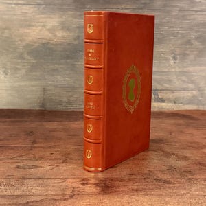 Puede incluir: Un libro antiguo de tapa dura con una cubierta naranja quemado. El lomo presenta dise&ntilde;os de tulipanes dorados y el t&iacute;tulo "SENSE & SENSIBILITY" y el autor "JANE AUSTEN". La portada tiene una silueta dorada enmarcada.