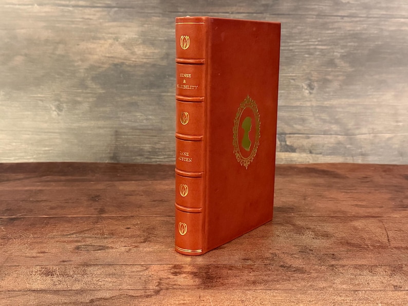 Puede incluir: Un libro antiguo de tapa dura con una cubierta naranja quemado. El lomo presenta dise&ntilde;os de tulipanes dorados y el t&iacute;tulo "SENSE & SENSIBILITY" y el autor "JANE AUSTEN". La portada tiene una silueta dorada enmarcada.