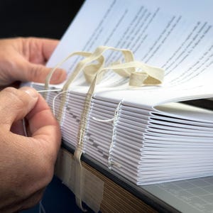 Puede incluir: Primer plano de un proceso de encuadernaci&oacute;n. Una pila de p&aacute;ginas de papel blanco se cose con hilo blanco y una aguja. Se utiliza cinta beige para asegurar las p&aacute;ginas. El libro est&aacute; sobre una esterilla de corte gris.