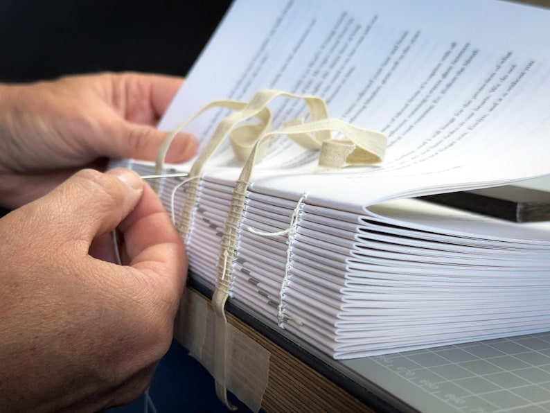 Puede incluir: Primer plano de un proceso de encuadernaci&oacute;n. Una pila de p&aacute;ginas de papel blanco se cose con hilo blanco y una aguja. Se utiliza cinta beige para asegurar las p&aacute;ginas. El libro est&aacute; sobre una esterilla de corte gris.
