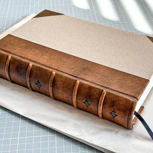 Puede incluir: Un diario encuadernado en cuero hecho a mano, con una cubierta beige y lomo de cuero marr&oacute;n. El lomo presenta detalles decorativos en relieve. Una cinta de marcador azul marino es visible.