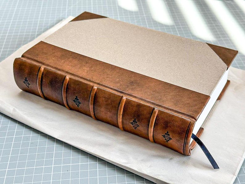Puede incluir: Un diario encuadernado en cuero hecho a mano, con una cubierta beige y lomo de cuero marr&oacute;n. El lomo presenta detalles decorativos en relieve. Una cinta de marcador azul marino es visible.