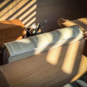 Puede incluir: Un objeto cil&iacute;ndrico de color claro con un detalle de cremallera, apoyado sobre una superficie de madera. El objeto est&aacute; parcialmente cubierto con una tela de color claro. Una caja marr&oacute;n y un recipiente con herramientas est&aacute;n en el fondo. La luz solar proyecta sombras.