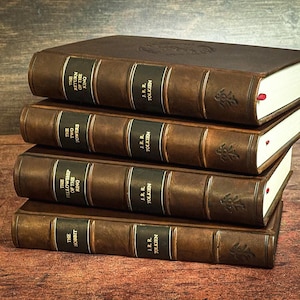 Puede incluir: Una pila de cuatro libros encuadernados en cuero marrón de J.R.R. Tolkien, incluyendo El Hobbit y la trilogía de El Señor de los Anillos. Cada lomo tiene etiquetas verde oscuro con letras doradas, nervios y un pequeño símbolo en relieve.