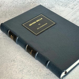 Puede incluir: Un libro de tapa dura azul oscuro titulado "SEVEN BELLS" de Roy Woodhouse. El libro tiene una placa de t&iacute;tulo dorada. El lomo presenta el t&iacute;tulo y el nombre del autor en letras doradas. El libro est&aacute; cerrado y sobre una superficie gris claro.