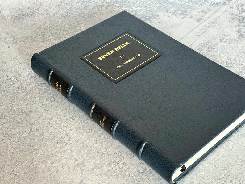 Puede incluir: Un libro de tapa dura azul oscuro titulado "SEVEN BELLS" de Roy Woodhouse. El libro tiene una placa de t&iacute;tulo dorada. El lomo presenta el t&iacute;tulo y el nombre del autor en letras doradas. El libro est&aacute; cerrado y sobre una superficie gris claro.