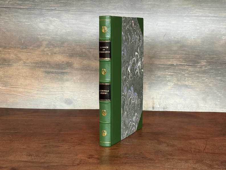 Puede incluir: Un libro de tapa dura con lomo verde y cubierta jaspeada. El lomo presenta letras doradas y elementos decorativos. El t&iacute;tulo del libro, "A Touch of Eternity" y el autor, "Isabella Gilbert" son visibles.
