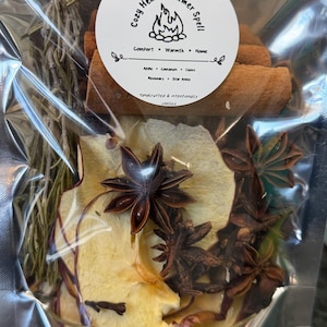 Op de afbeelding: Een doorzichtige plastic zak met gedroogde ingrediënten voor een sudderpot, gelabeld 'Shaman Tiff Cozy Hearth Simmer Spell'. De inhoud bestaat uit gedroogde appelschijfjes, kaneelstokjes, steranijs, hele kruidnagels en een bundel gedroogde rozemarijntakjes.