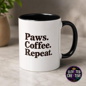 May include: A white ceramic mug with a black interior and handle. The exterior features the dark brown text Paws. Coffee. Repeat. printed in a bold, sans-serif font. A circular logo with the text ELEVATED CRE8TIVE Premium Tees is visible.