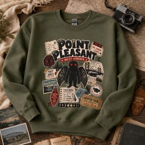 Può includere: Una felpa girocollo color verde oliva con una stampa grafica del Mothman dagli occhi rossi, circondata da illustrazioni in stile vintage che includono il testo Point Pleasant West Virginia, un ponte, appunti sul campo e vari elementi grafici.
