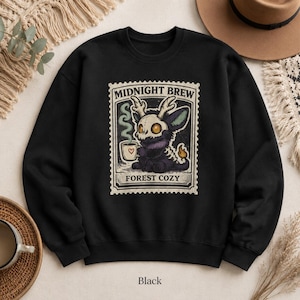 May include: A black long-sleeve sweatshirt featuring a graphic print of a creature with antlers and a skull-like face holding a steaming mug. The design is framed like a postage stamp with the text MIDNIGHT BREW and FOREST COZY printed in black.