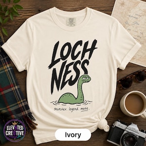 May include: An ivory short-sleeve t-shirt featuring a black graphic of the Loch Ness monster with the text "LOCH NESS" in a bold, brush-stroke font above it and the text "monster. legend. mate." in a smaller, simple font below the creature.