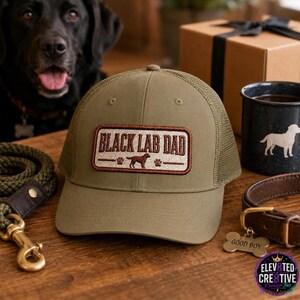 May include: An olive green trucker hat featuring a rectangular patch with brown embroidered text that reads BLACK LAB DAD, accompanied by a small dog silhouette and paw prints. The cap has a solid front panel and a mesh back for ventilation.