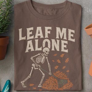 May include: A brown t-shirt with the words "LEAF ME ALONE" in white. The design shows a skeleton raking leaves, a humorous take on autumn and gardening.