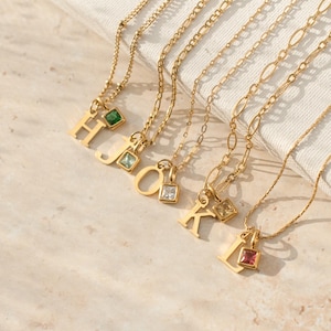May include: Gold-colored necklaces with initial pendants. Each necklace features a different letter: H, J, O, K, and L. The pendants are adorned with colorful gemstones, including green, blue, clear, yellow, and red. The necklaces are displayed on a white surface.