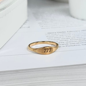 以下が含まれることがあります： 上部に777が刻まれた金のシグネットリング。リングは本の上に置かれています。