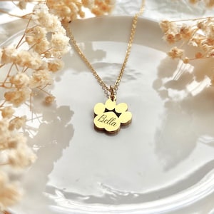 May include: Gold paw print necklace with the name "Bella" engraved on it. The necklace is on a white plate with white flowers in the background.