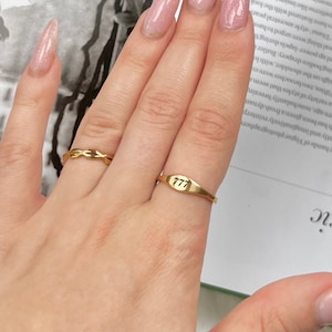 以下が含まれることがあります： 2つの金の指輪をした手。人差し指の指輪は、ねじれたデザインの細いバンド。中指の指輪は、太いバンドで、数字の「777」が刻まれている。