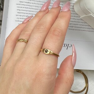 May include: A gold ring featuring a black paw print design. The ring is worn on a person's finger.