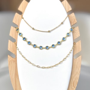Può includere: Tre collane d'oro su un espositore bianco. La collana superiore è una semplice catena con piccole perle dorate. La collana centrale ha una fila di perle di pietre preziose blu. La collana inferiore è una catena delicata.