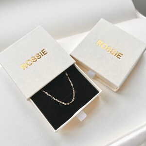 May include: Two white boxes with the word "ROSSIE" printed in gold lettering. One box is open and contains a gold chain necklace on a black velvet background.