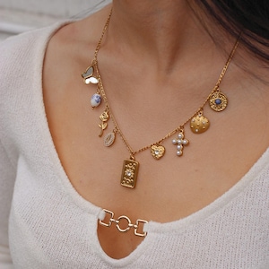 K&ouml;nnte beinhalten: Eine goldfarbene Kettenhalskette mit mehreren Anh&auml;ngern, darunter ein Schmetterling, eine blau-wei&szlig; gemusterte Perle, eine Rose, eine Muschel, ein rechteckiger Anh&auml;nger mit Sonnendesign, ein Herz, ein Kreuz mit Perlen und ein rundes Auge-Symbol.