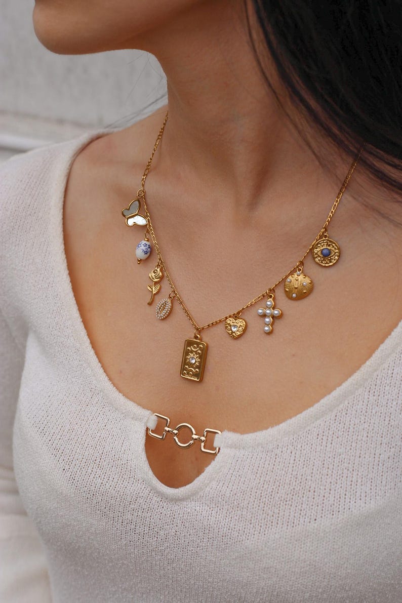 K&ouml;nnte beinhalten: Eine goldfarbene Kettenhalskette mit mehreren Anh&auml;ngern, darunter ein Schmetterling, eine blau-wei&szlig; gemusterte Perle, eine Rose, eine Muschel, ein rechteckiger Anh&auml;nger mit Sonnendesign, ein Herz, ein Kreuz mit Perlen und ein rundes Auge-Symbol.