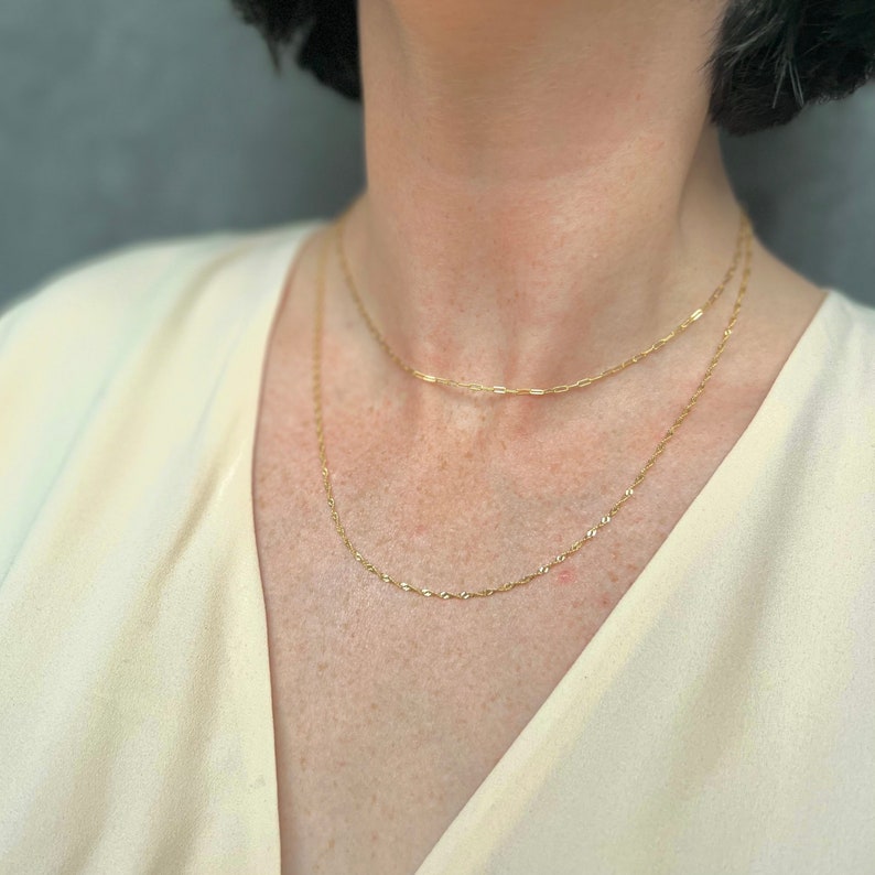 May include: Two gold necklaces layered on a person's neck. The top necklace is a thin chain with small, rectangular links. The bottom necklace is a slightly thicker chain with oval links.