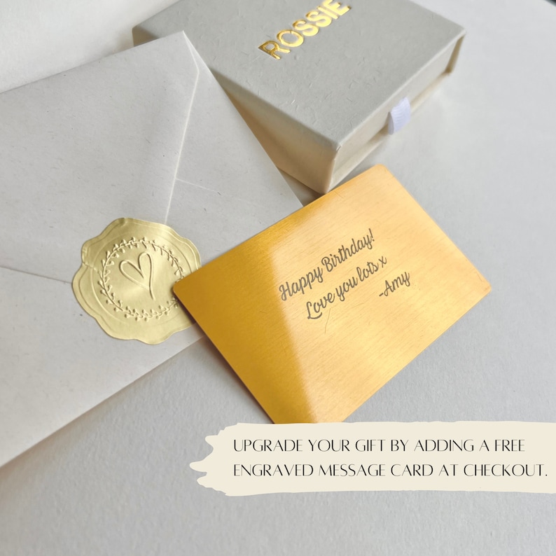 K&ouml;nnte beinhalten: Eine goldfarbene Karte mit einer handgeschriebenen Nachricht, die lautet "Happy Birthday! Love you lots x -Amy". Die Karte liegt auf einer wei&szlig;en Oberfl&auml;che mit einem wei&szlig;en Umschlag und einer kleinen goldfarbenen Schachtel, auf der das Wort "ROSSIE" steht.