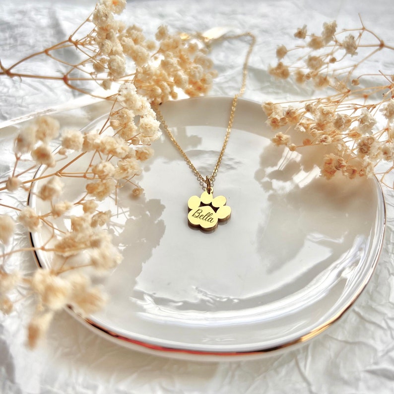 May include: Gold paw print necklace with the name "Bella" engraved on it. The necklace is on a white plate with white flowers in the background.