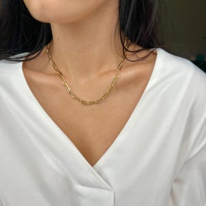 K&ouml;nnte beinhalten: Eine goldene Kettenkette mit einem einfachen, verlinkten Design. Die Kette wird von einer Person mit dunklen Haaren und einem wei&szlig;en Hemd getragen.