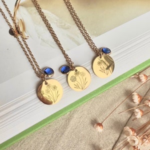 Könnte beinhalten: Drei goldene Halsketten mit zarten Blumenmotiven und blauen Edelstein-Akzenten. Jede Halskette hat einen runden Anhänger mit einem anderen Blumenmotiv.