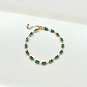 Op de afbeelding: Een gouden ketting met rechthoekige smaragdgroene stenen. De ketting is delicaat en heeft een eenvoudig ontwerp.