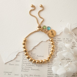 Pode incluir: Um bracelete de contas douradas com um fecho deslizante dourado e um pequeno amuleto dourado com o nome "Ava" nele. O bracelete também tem três pequenas pedras preciosas coloridas presas ao amuleto.
