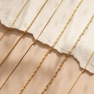 May include: A collection of gold chains of different styles and thicknesses laid out on a beige surface.