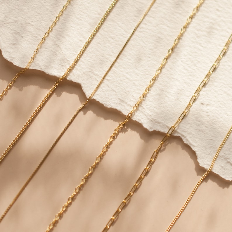 May include: A collection of gold chains of different styles and thicknesses laid out on a beige surface.