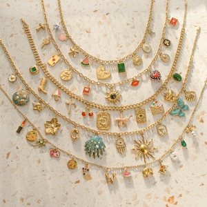K&ouml;nnte beinhalten: Eine Sammlung von f&uuml;nf goldfarbenen Ketten, die jeweils mit einer Auswahl an bunten Emaille- und Metallanh&auml;ngern verziert sind, darunter Symbole wie Sonnen, Muscheln, Herzen, Sterne, Fr&uuml;chte, Buchstaben und verschiedene andere Motive.