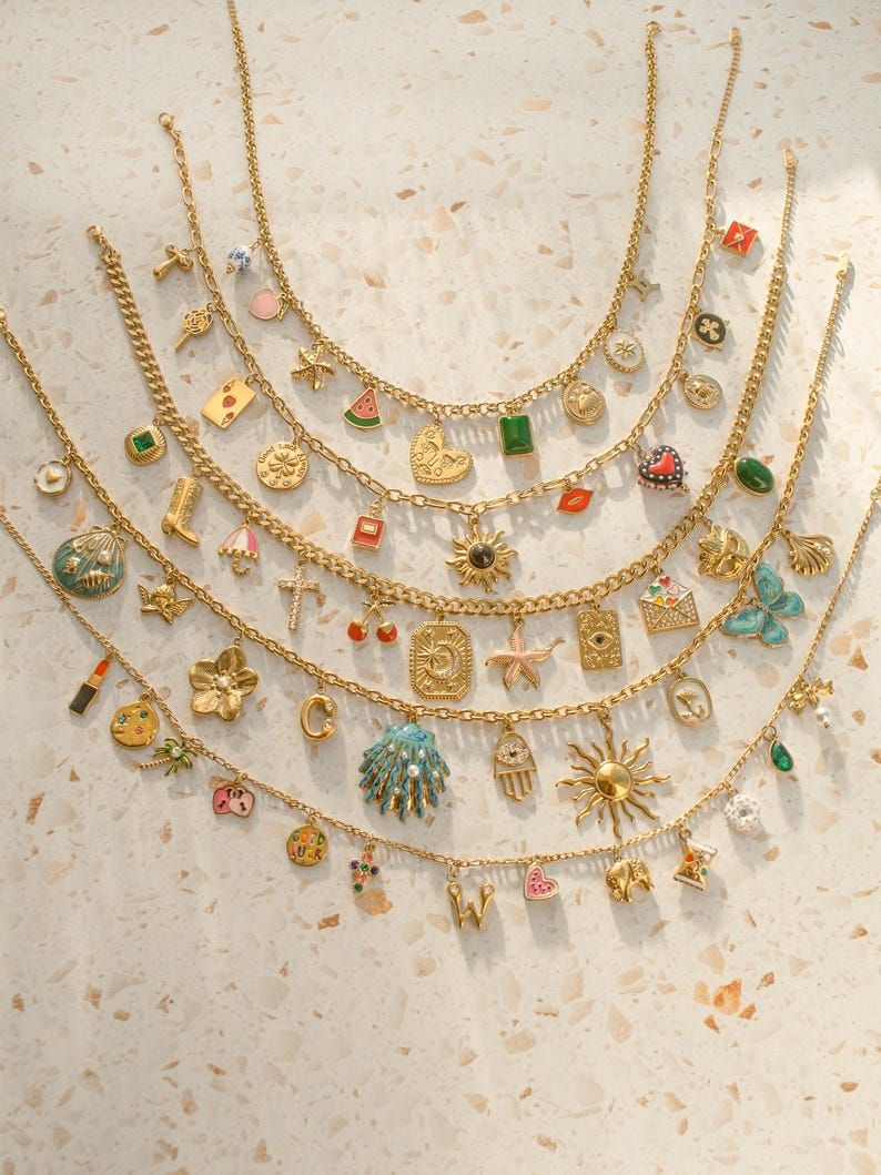 K&ouml;nnte beinhalten: Eine Sammlung von f&uuml;nf goldfarbenen Ketten, die jeweils mit einer Auswahl an bunten Emaille- und Metallanh&auml;ngern verziert sind, darunter Symbole wie Sonnen, Muscheln, Herzen, Sterne, Fr&uuml;chte, Buchstaben und verschiedene andere Motive.
