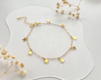 Puede incluir: Una delicada pulsera dorada con pequeños abalorios en forma de estrella. La pulsera se muestra sobre una superficie clara y ondulada, con flores secas en el fondo. La cadena de la pulsera es fina y las estrellas están espaciadas uniformemente.