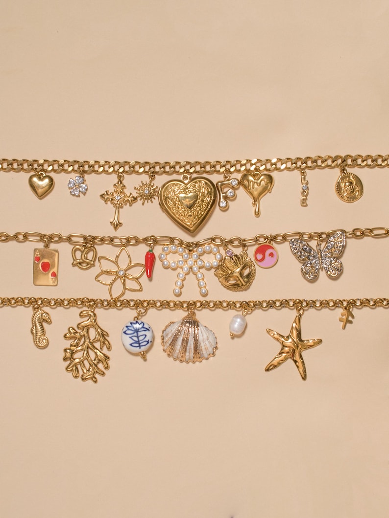 K&ouml;nnte beinhalten: Drei goldfarbene Bettelarmb&auml;nder mit einer Vielzahl von Anh&auml;ngern, darunter ein Herzmedaillon, ein Kreuz, eine mit Perlen besetzte Schleife, ein Schmetterling, eine Muschel, ein Seestern, ein Seepferdchen, eine Spielkarte und eine Chilischote.