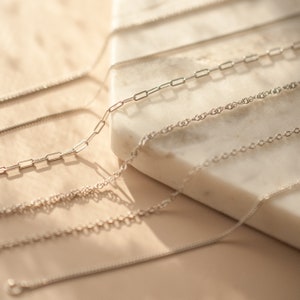 May include: A close-up of five silver chains of different styles and thicknesses, laid out on a white marble surface.